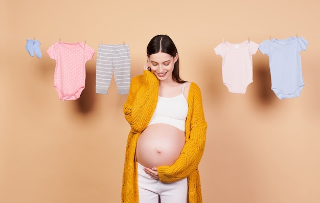 A nice, smiling young girl with long dark hair, in a yellow knitted cardigan, with a bare belly, touches her belly with her hands and looks at him with a sweet smile. Motherhood, childhood concept.