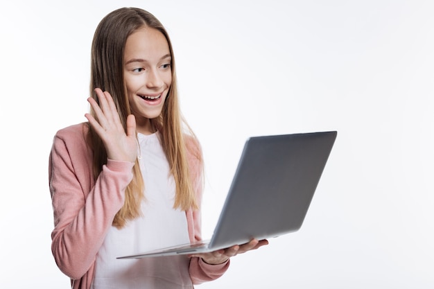 Рад вас видеть. очаровательная девочка-подросток держит ноутбук и делает видеозвонок, машет перед веб-камерой и улыбается