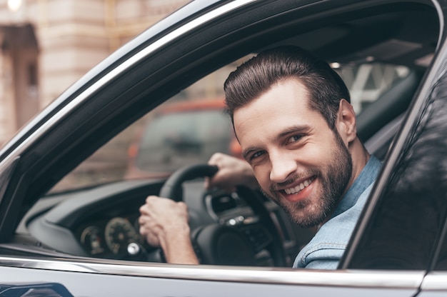 Nice travel. Confident young man smiling and looking at camera while driving a car