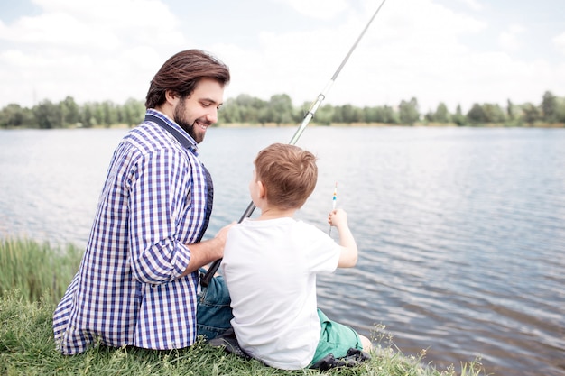Nice view of happy son and dad sitting together at river shore. Guy is looking at his son and fishing. Boy is looking at his father and talking to him.