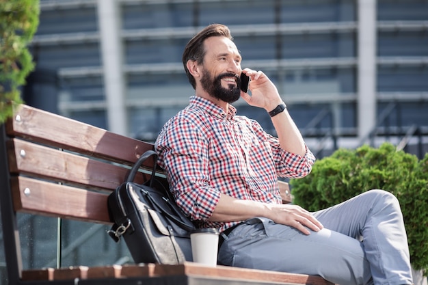 Nice weather. Cheerful pleasant man resting on the bench while enjoying phone conversation