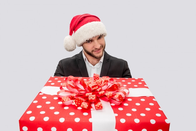 Nice young man looks on camera and holds big red box with present. He wears suit and Christmas hat. 