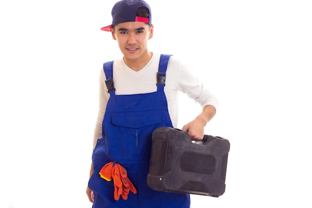 Nice young man with dark hair in white shirt and blue overall with orange gloves and blue snapback holding toolbox on white background in studio