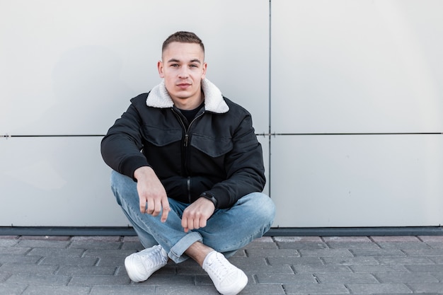 Nice young man with a stylish hairstyle in a fashionable black jacket in vintage jeans in leather trendy sneakers resting sitting on a gray tile near a modern wall on a bright spring day