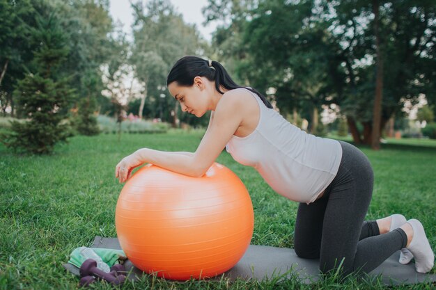 Nice young pregnant woman having workout in park. She stand on knees and look down. Model lean to big orange fitness ball.