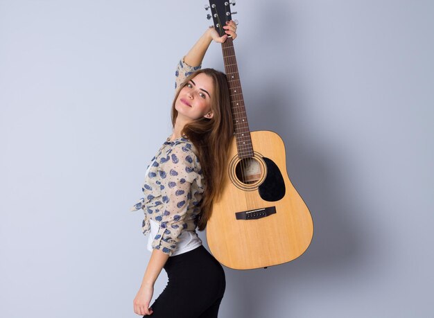 Nice young woman in beige blouse and black trousers holding a guitar on gray background in studio