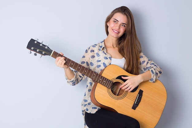 Nice young woman in beige blouse and black trousers playing the  guitar on gray background in studio