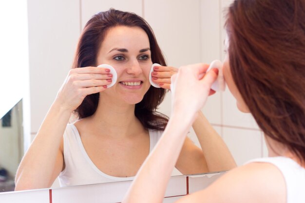 Nice young woman in shirt removing makeup with cotton pad in front of the mirror in her bathroom