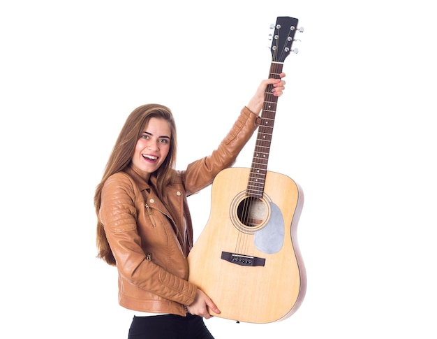 Nice young woman with long hair in brown jacket and black trousers holding a guitar on white background in studio