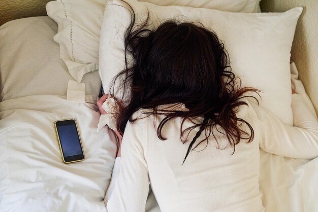 Niemand begrijpt je beter dan je bed Hoge hoekopname van een jonge vrouw die in bed ligt met tissues en haar mobiele telefoon naast haar