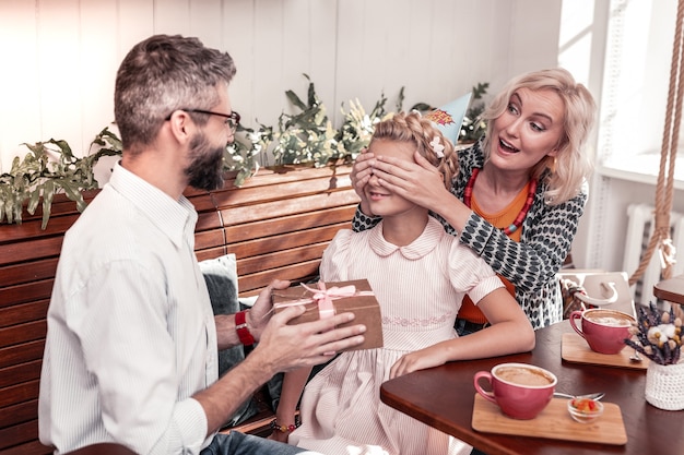 Niet kijken. aangename jonge vrouw die de ogen van haar dochters bedekt terwijl ze een cadeau voor haar klaarmaakt