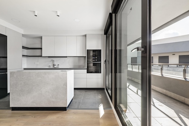 Nieuw geplaatste moderne keuken met centraal eiland gladde grijze kasten zwart glazen apparaten en glazen wand met toegang tot een open terras