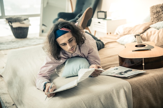 Nieuwe compositie maken. doordachte jongeman in kleurrijke bandana die notities in een speciaal album observeert terwijl hij op de buik in de slaapkamer ligt