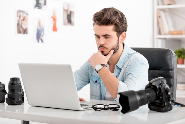 Nieuwe ideeën genereren. Geconcentreerde jonge man die hand op kin houdt en naar laptop kijkt terwijl hij op zijn werkplek zit