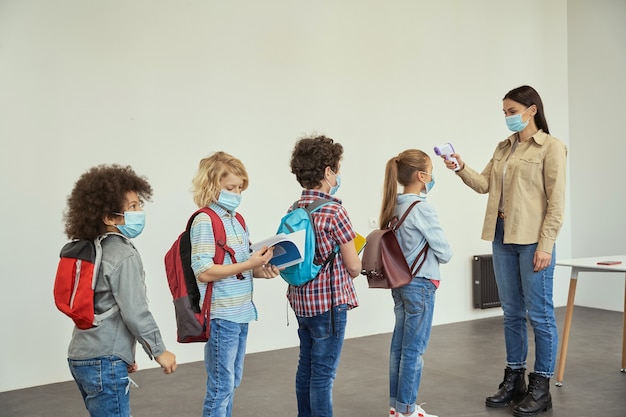 Nieuwe regels moderne jonge vrouwelijke leraar die temperatuurscreening van kinderen meet met digitale thermometer