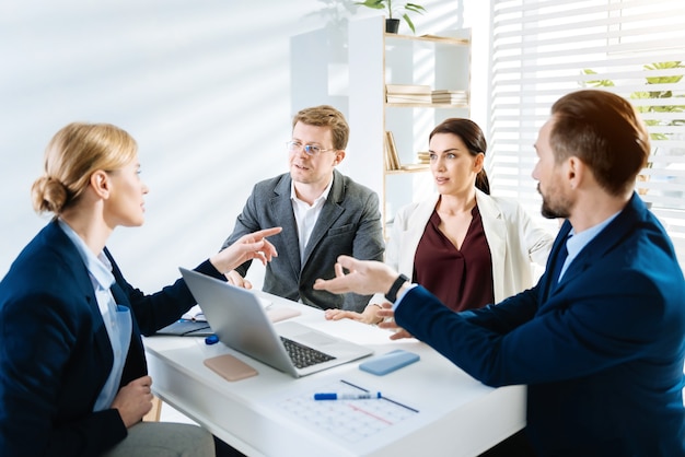 Nieuwe zakelijke stijl. Verrast jonge vrouwelijke collega nieuws vertellen terwijl haar collega's aan tafel zitten en luisteren