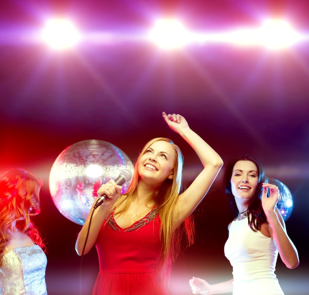 nieuwjaar, feest, vrienden, vrijgezellenfeest, verjaardagsconcept - drie vrouwen in avondjurken dansen en zingen karaoke