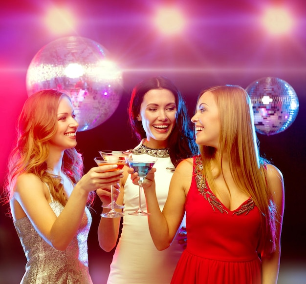 nieuwjaar, feest, vrienden, vrijgezellenfeest, verjaardagsconcept - drie vrouwen in avondjurken met cocktails en discobal