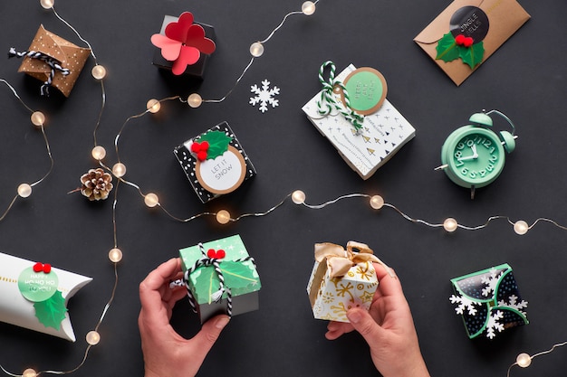 Nieuwjaar of kerstcadeautjes verpakt in verschillende papieren geschenkdozen met feestelijke tags. twee handen met vakken. feestelijke plat lag, bovenaanzicht met lichte garland, wekker en sneeuwvlokken op zwart papier.