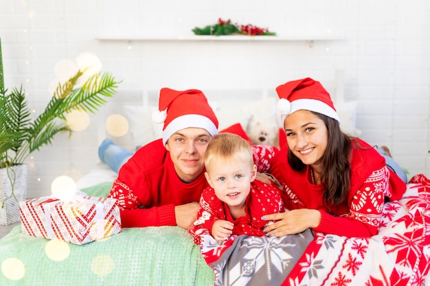 Nieuwjaar of Kerstmis, een gelukkig gezin met een kind op het bed thuis bij de kerstboom in rode truien glimlachen, knuffelen en zoenen feliciteren met de vakantie