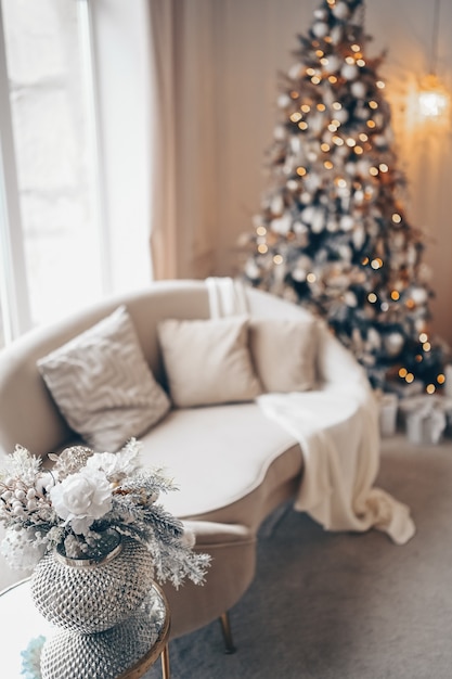 Nieuwjaars vakantie decoratie boeket in zilveren vaas op nachtkastje glazen tafel tegen witte sofa en versierde kerstboom met garland lichten