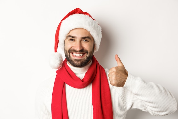 Nieuwjaarsfeest en wintervakantie concept. Close-up van een gelukkige man in een kerstmuts die zijn duimen omhoog laat zien, leuk vindt en akkoord gaat, staande op een witte achtergrond