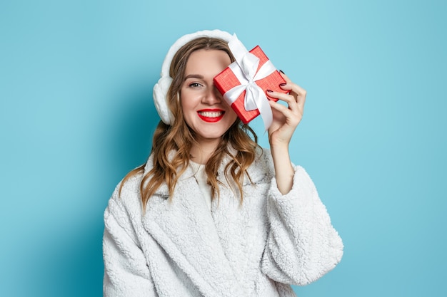 Nieuwjaarsgeschenken 2021. jonge gelukkig lachende vrouw in een witte kunstbontjas en bont koptelefoon sluit haar ogen met een geschenkdoos en glimlacht geïsoleerd op een blauwe muur. kopieer ruimte