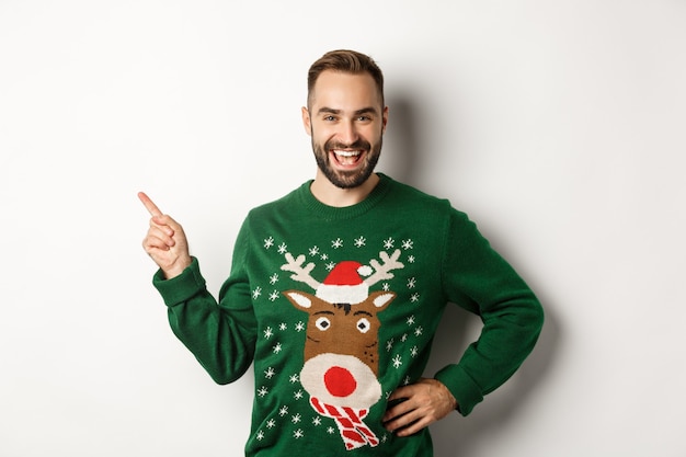 Nieuwjaarsviering en wintervakantie concept. Knap bebaard mannelijk model in kersttrui lachen, wijzende vinger naar het logo van de linkerbovenhoek, witte achtergrond.