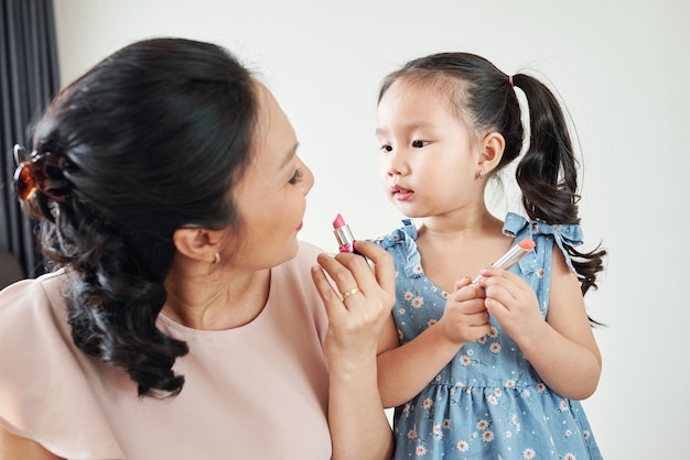 Nieuwsgierig schattig klein Vietnamees meisje kijkt naar haar moeder die lichtroze lippenstift toepast wanneer ze zich 's ochtends klaarmaakt