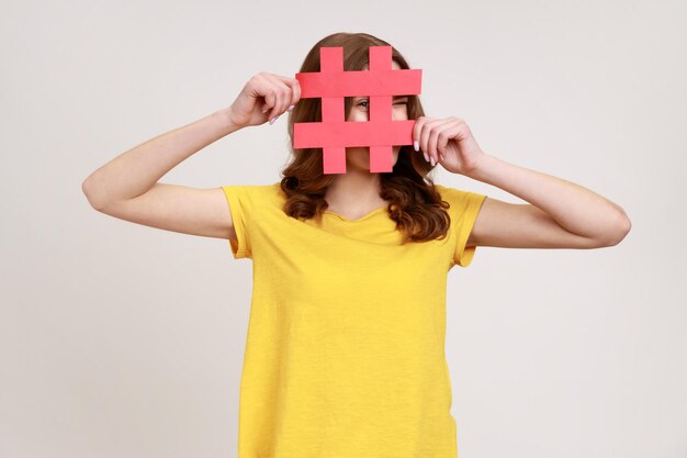 Nieuwsgierige bruinharige vrouw van jonge leeftijd in geel T-shirt die door een rood hashtag-teken kijkt, op zoek naar goede berichten op sociale media, spionage Indoor studio opname geïsoleerd op een grijze achtergrond.
