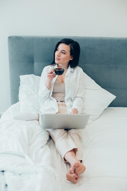 Nieuwsgierige dame met een kopje koffie in de verte kijkend zittend op een bed met een laptop op haar benen