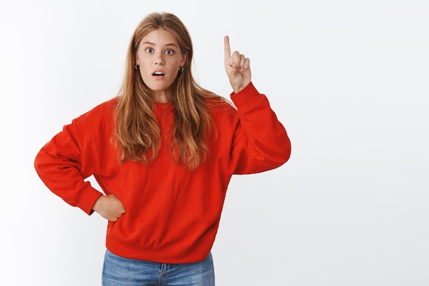 Nieuwsgierige en geïntrigeerde aantrekkelijke jonge mollige vrouw met rood haar, sproeten en blauwe ogen in warme trendy rode trui met open mond ondervraagd hand opsteken en naar boven wijzend, vraag stellend