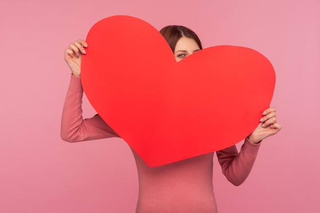 Nieuwsgierige grappige vrouw in roze trui die zich verschuilt achter een groot papieren hart, heilige valentijnsdagviering. Indoor studio-opname geïsoleerd op roze background