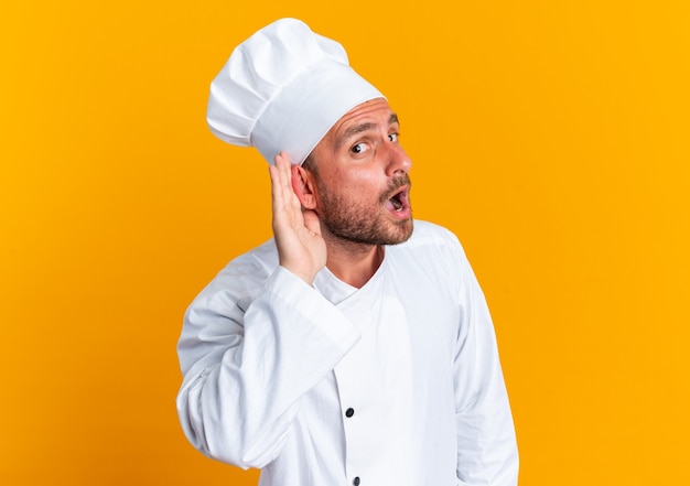 Nieuwsgierige jonge blanke mannelijke kok in uniform van de chef-kok en pet kijkend naar de camera, ik hoor je gebaar niet geïsoleerd op een oranje muur