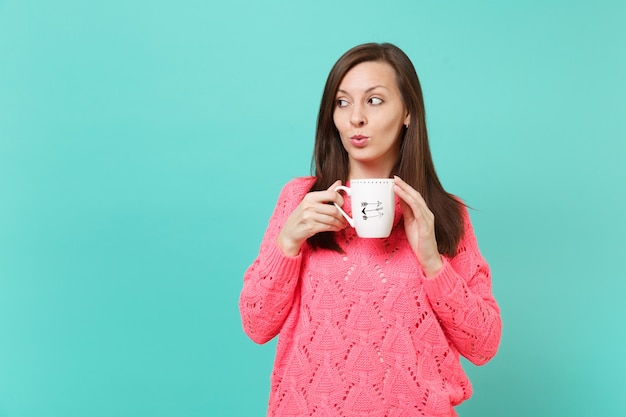 Nieuwsgierige jonge vrouw in gebreide roze trui opzij kijken, in de hand kopje koffie of thee houden geïsoleerd op blauwe turquoise muur achtergrond, studio portret. Mensen levensstijl concept. Bespotten kopie ruimte.