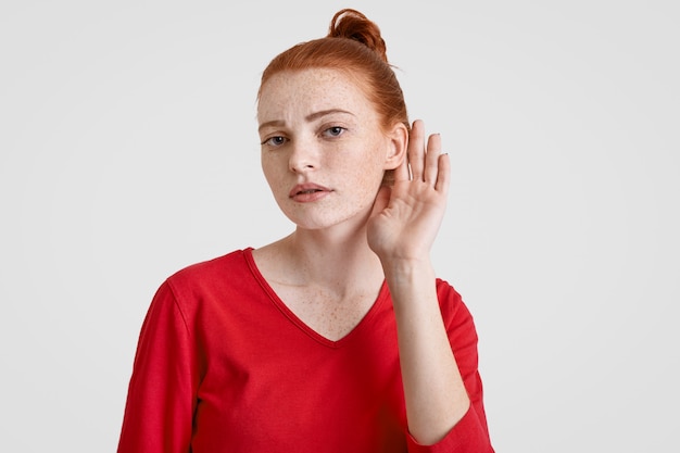 Nieuwsgierige jonge vrouw met sproeten met gemberbroodje houdt haar hand in de buurt van het oor, probeert vertrouwelijke informatie te horen, houdt van roddelen, gekleed in een rode casual trui, geïsoleerd op een witte studio.