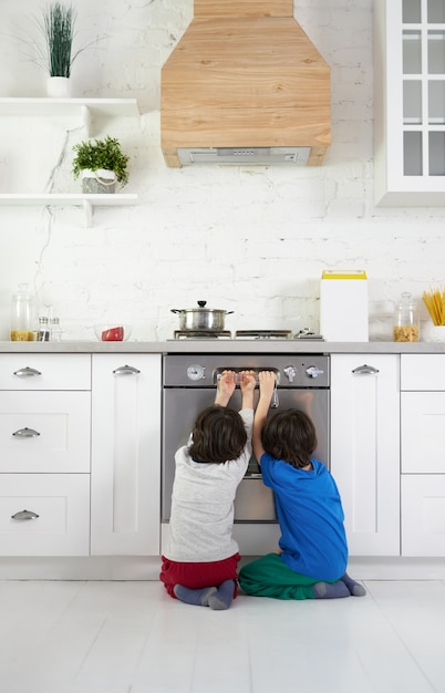 Nieuwsgierige Latijns-Amerikaanse jongens, tweeling kijken naar taart bakken in de oven, gehurkt in de keuken thuis. Kinderen, kookconcept. Achteraanzicht