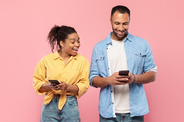 Nieuwsgierige zwarte vrouw die probeert te kijken naar de gsm-man van haar vriend die zijn gadget gebruikt en op een roze achtergrond kletst