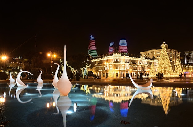 Ночной Баку в Новом году со светящейся елкой, украшенной узорами, и фонтаном-лебедем на переднем плане.