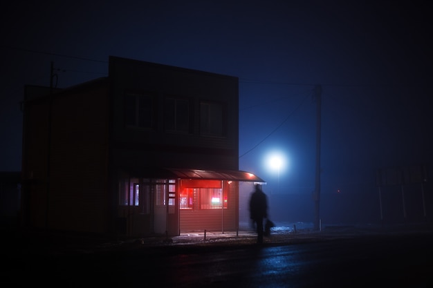 夜の青い霧の風景。赤く照らされた看板のある小さなお店が道路の脇に立っており、脂っこい男のシルエットが道路を横切っています。