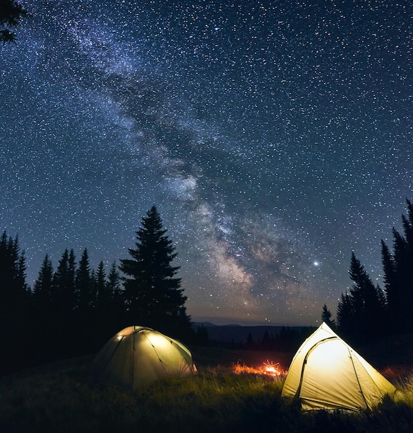 天の川がはっきりと見える明るい星空の下、松林に焚き火と2つのテントが燃えるナイトキャンプ遠くに丘や山々の谷が見える強大な風景