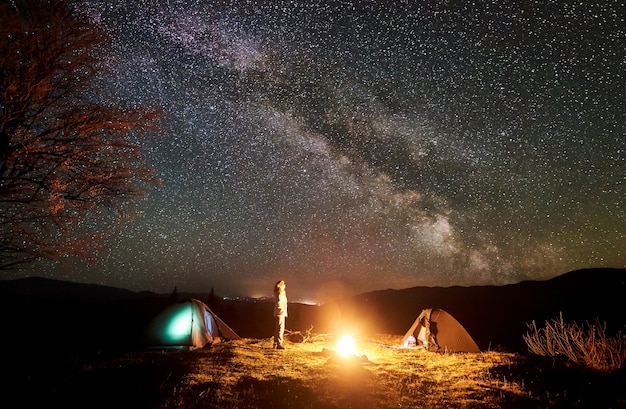 Night camping. Hiker resting near campfire under starry sky