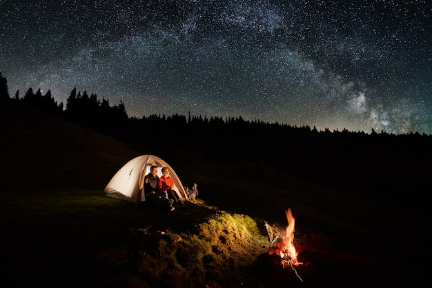 写真 山でのキャンプ。カップルの観光客は、星と天の川でいっぱいの美しい夜空の下でキャンプファイヤーの近くの照らされたテントで残りを持っています