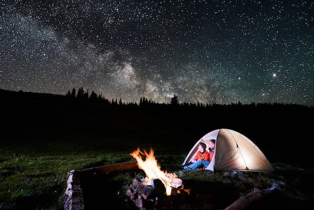 Night camping in the mountains. Romantic couple tourists sitting in the illuminated tent near campfire and looking at amazing night sky full of stars and milky way