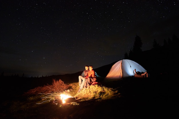 夜のキャンプ。ロマンチックなカップルの観光客は夜の星空の下で照らされたテントの近くのキャンプファイヤーで残りを持っています