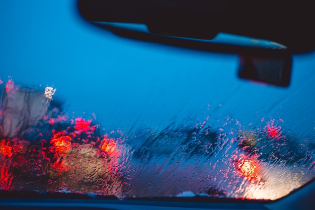 La strada della città di notte attraverso le automobili del parabrezza astraggono la goccia d'acqua del fondo sulle luci e sulla pioggia di vetro.