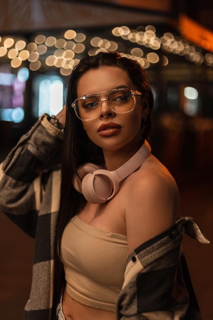 Night female portrait of a beautiful stylish woman with fashion eyewear glasses and pink earphones in trendy street clothes walks in the night city with bokeh lights