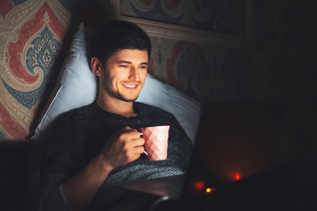 Night  young cheerful man lying on bed in dark room at home with cup of coffee and laptop.