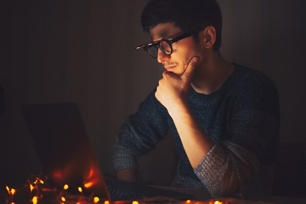 Giovane uomo premuroso di notte con gli occhiali rotondi, guardando nel computer portatile in camera oscura con ghirlande a casa.