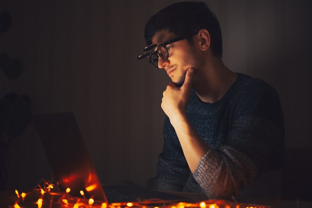 Night  young thoughtful man wearing round glasses, looking in laptop in dark room with garlands at home.